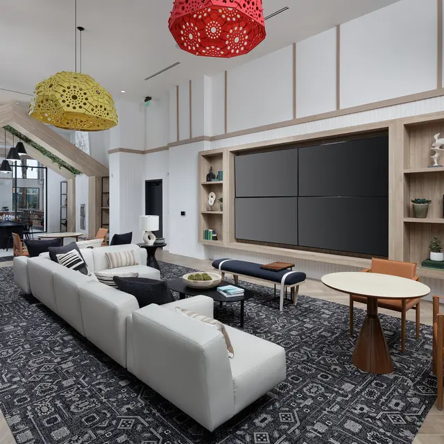 A modern living room featuring a large white couch, colorful hanging lamps, a stylish area rug, a wooden shelf with decorative items, and a wall-mounted television.
