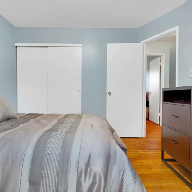 A cozy bedroom featuring a bed with a striped gray and silver comforter. The walls are painted light blue, and there is a wooden dresser with a decorative frame and candles on top. White closet doors are visible opposite the bed, and a doorway leads to another room in the background.