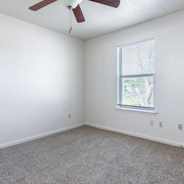 An empty room with light gray carpet and a ceiling fan. There is a window with blinds facing outside, and the walls are painted white.