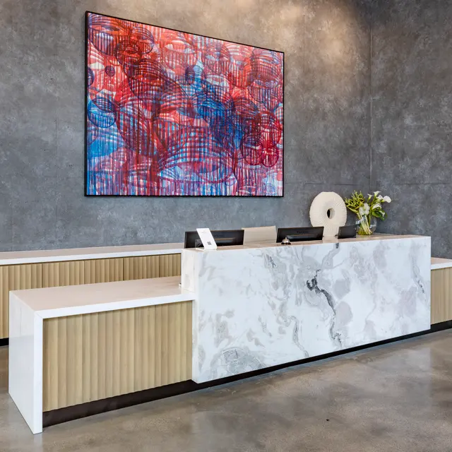 A modern reception area featuring a sleek front desk with a marble base and wooden accents, accompanied by a colorful abstract artwork on the wall. The space has a polished concrete floor and a contemporary design.