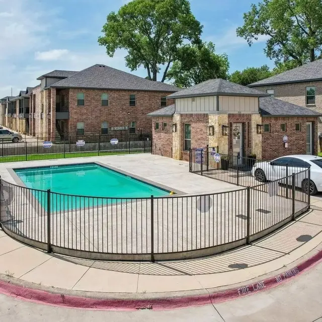 A view of an apartment complex featuring a swimming pool surrounded by a black fence. The pool is rectangular, and there are parked cars nearby. Two buildings are visible in the background with several trees.