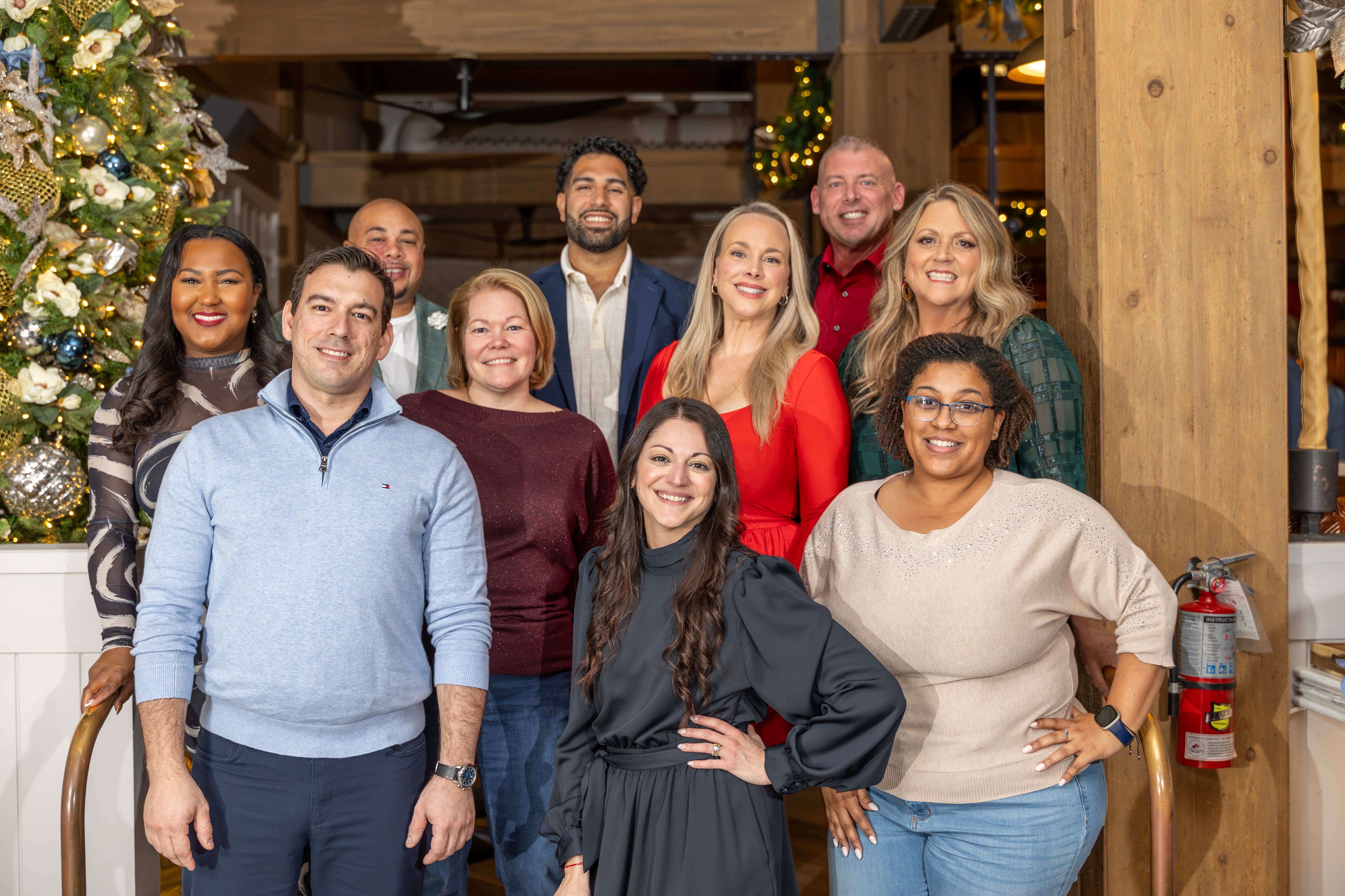 A festive group photo featuring a diverse group of eleven individuals, posing together inside a decorated venue, with holiday ornaments and trees in the background.