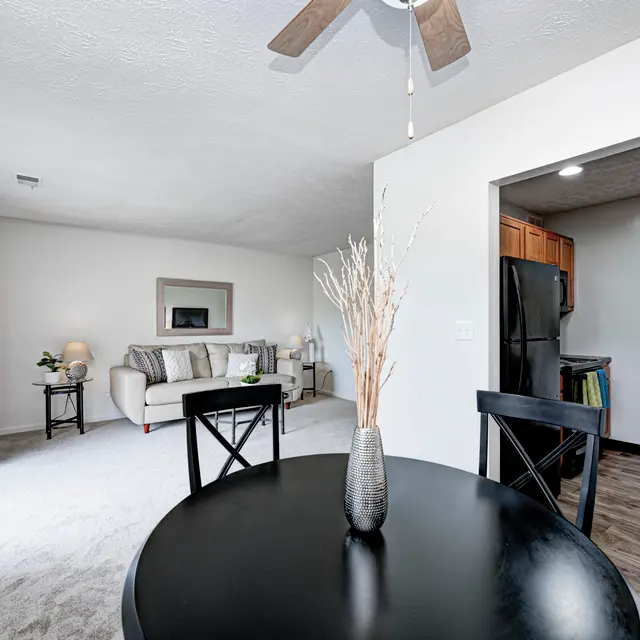 A modern apartment interior featuring a dining table with a vase, a living room area with a sofa and lamp, and a kitchen visible in the background.