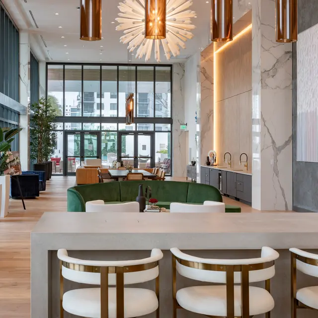 A modern and stylish lobby interior featuring large windows, modern furniture, and decorative plants. The space includes a bar area with white stools, a green sofa, and unique lighting fixtures.