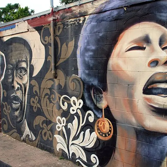 A vibrant mural featuring three expressive human faces - a man with sunglasses, a serious-looking man, and a woman singing passionately. The artistic style combines realism with stylized floral patterns, adding depth to the scene.