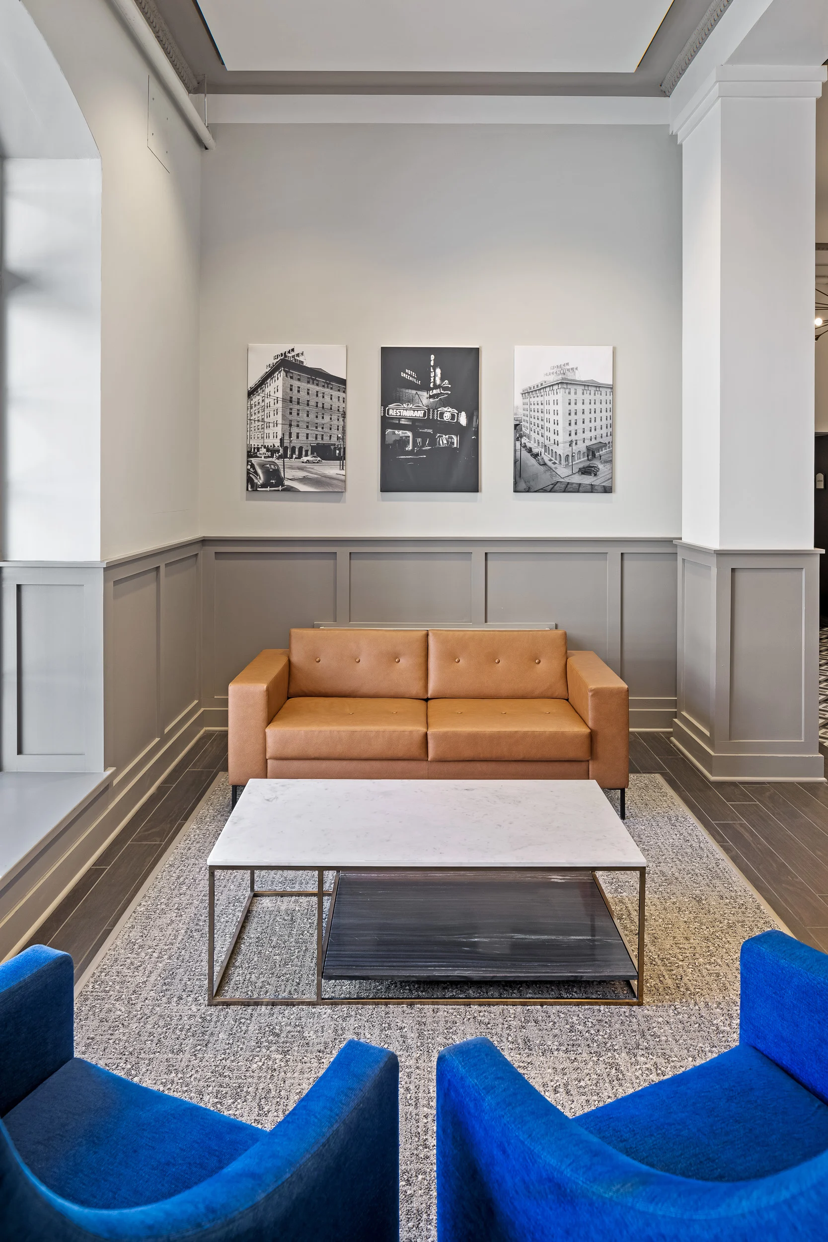 A cozy modern lounge area featuring a brown leather sofa, a square coffee table, and two blue armchairs. The walls are light-colored with framed black and white photographs. There is a patterned rug on the floor.