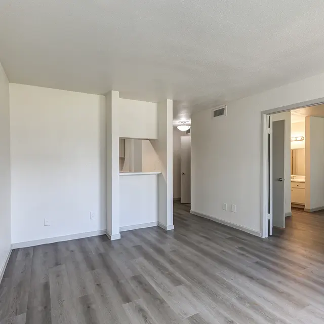 A modern apartment interior featuring light gray walls, laminate flooring, and an open floor plan. The left side has a sunlit kitchen area with a partial wall, and a doorway leading to another room is visible on the right.
