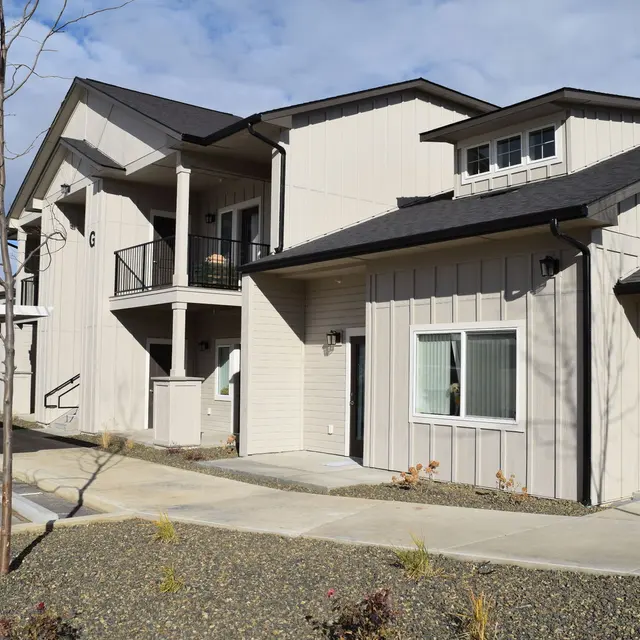 Modern Apartment Building A modern two-story apartment building with beige siding and black roofing. The scene includes a landscaped area with gravel, small plants, and a paved walkway leading up to the units.