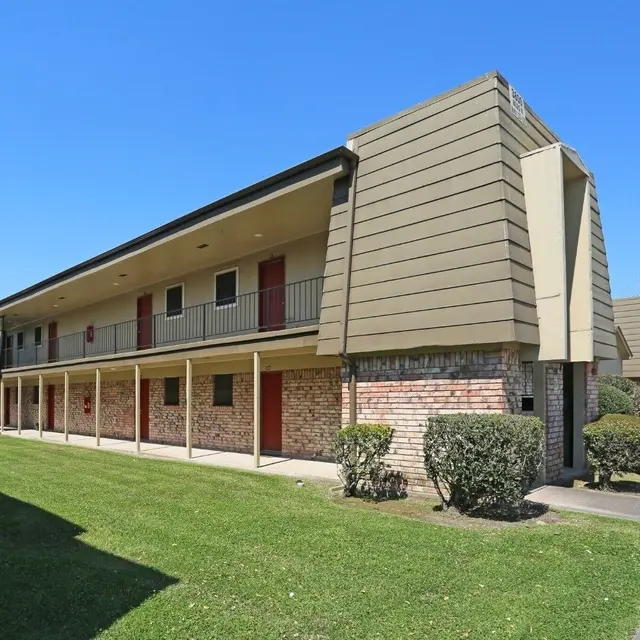 Exterior view of a two-story apartment building with a green lawn and shrubs in front, under a clear blue sky.