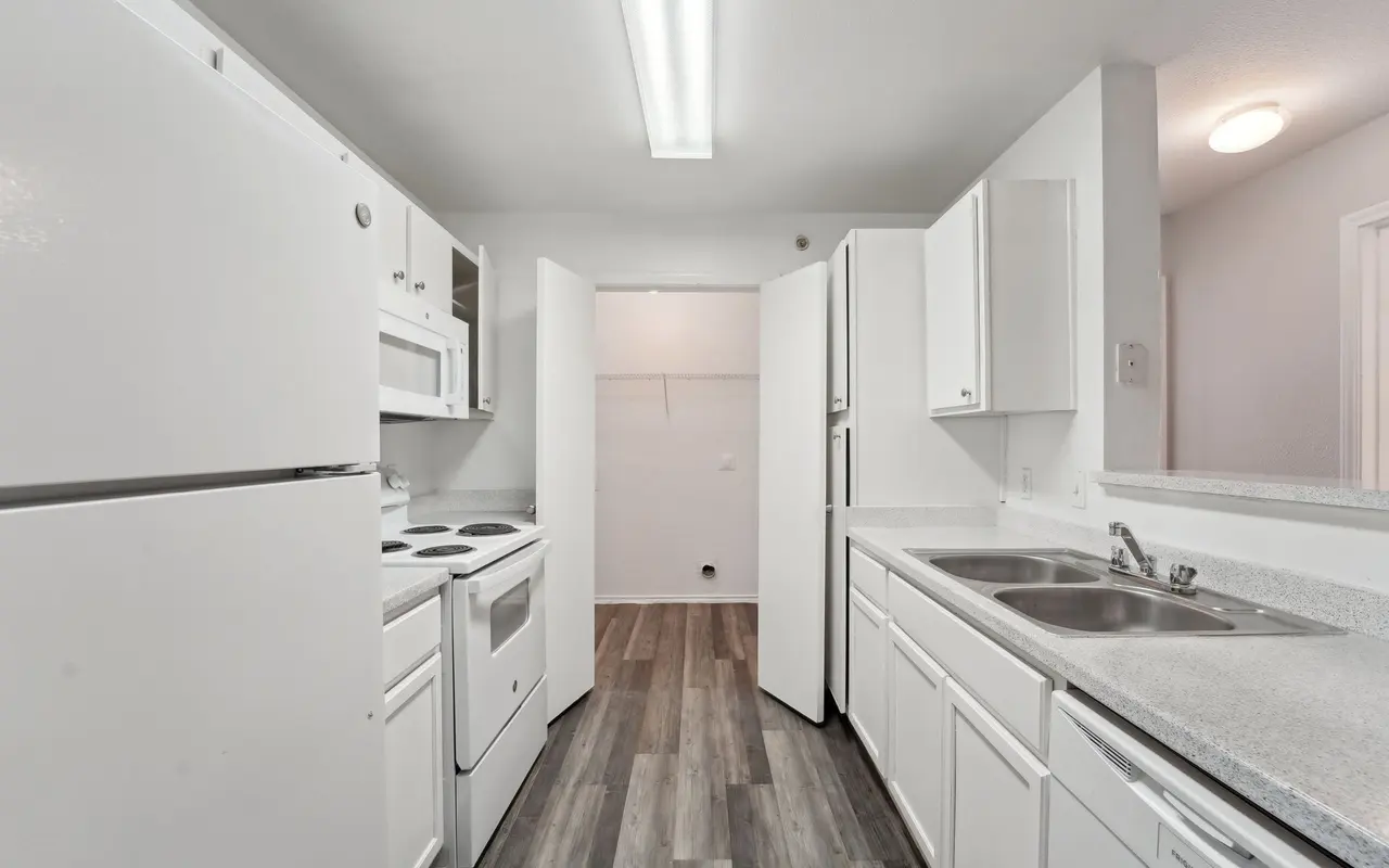 A modern kitchen featuring white cabinetry, appliances, and a light-colored countertop. The space has a clean and minimalistic design with a floor made of planks. Two doors lead to additional areas.