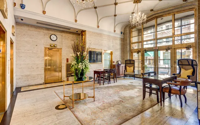 A spacious hotel lobby featuring elegant furniture and large windows. The floor is adorned with a decorative rug, and there are chairs and a table arranged neatly. An elevator is visible in the background, along with a clock and decorative flower arrangements.