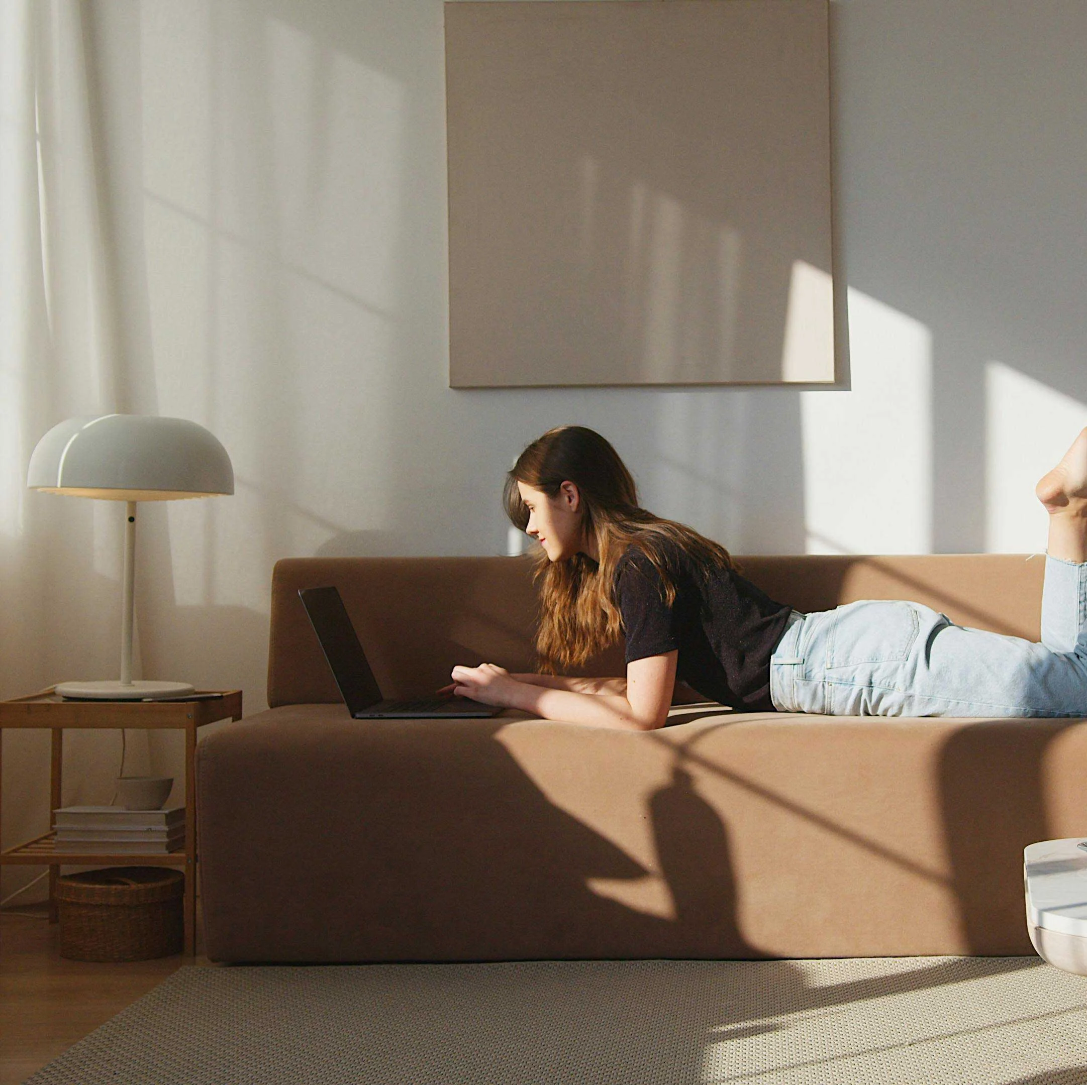 A young woman lying on a couch, working on a laptop in a well-lit room with a lamp and a small table beside her.