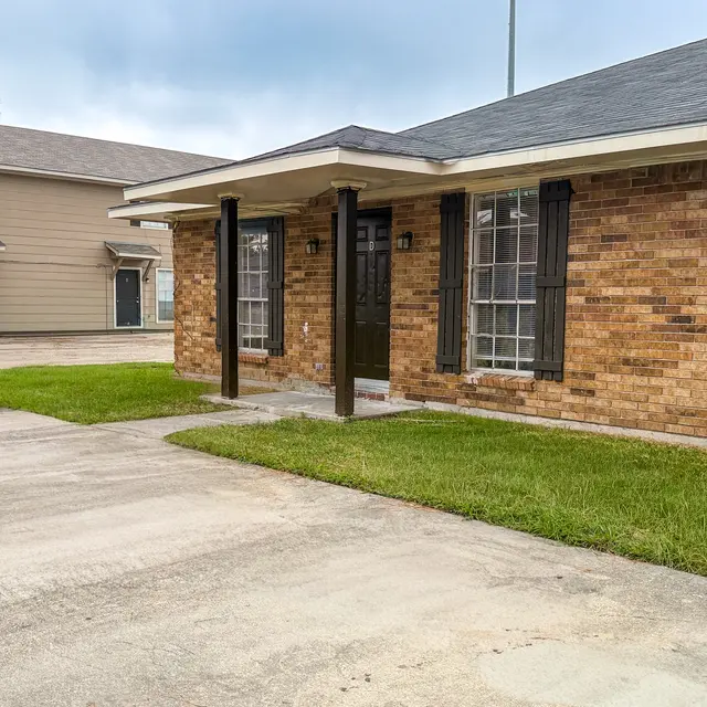 A brick house featuring a wide front porch, with green grass and a concrete driveway in the foreground. A second building can be seen in the background, adding to the residential setting.