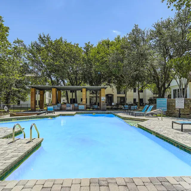 A clear swimming pool surrounded by green trees and lounge chairs.