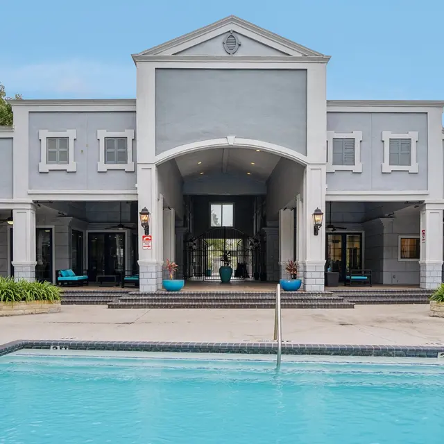 A modern residential complex with a large pool in front and a spacious building in the background. The building features a grand entrance and is surrounded by greenery and decorative planters. Blue lounge chairs are placed around the pool area.