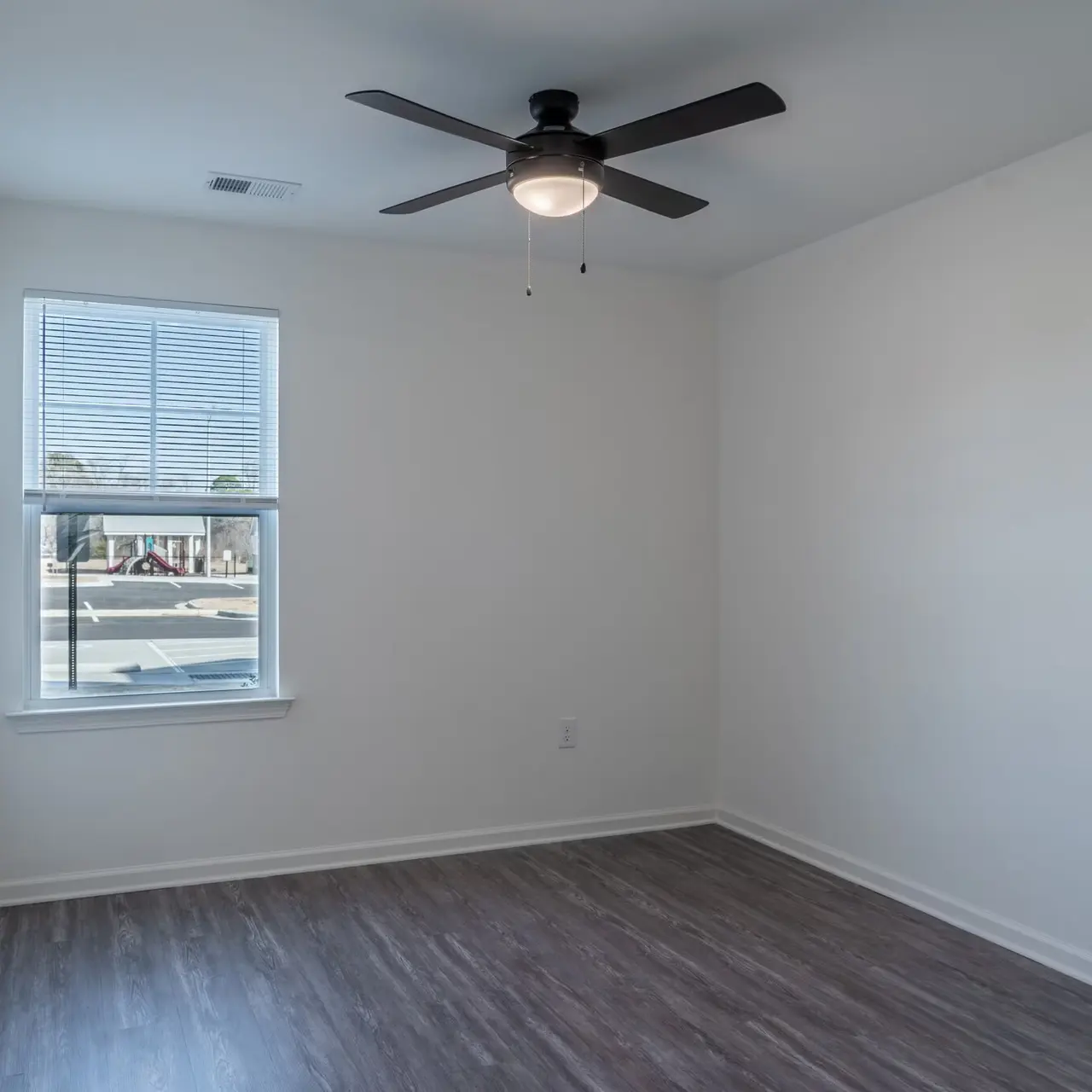 An empty room with a ceiling fan and a window showing outside. The walls are painted white and the floor has a dark laminate finish.