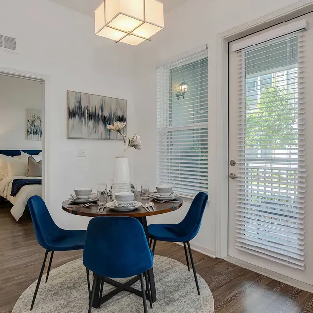 A stylish dining area featuring a round table with blue chairs, set for a meal, adjacent to a room with a bed visible through a doorway. The space is well-lit by a modern chandelier and has large windows with blinds.