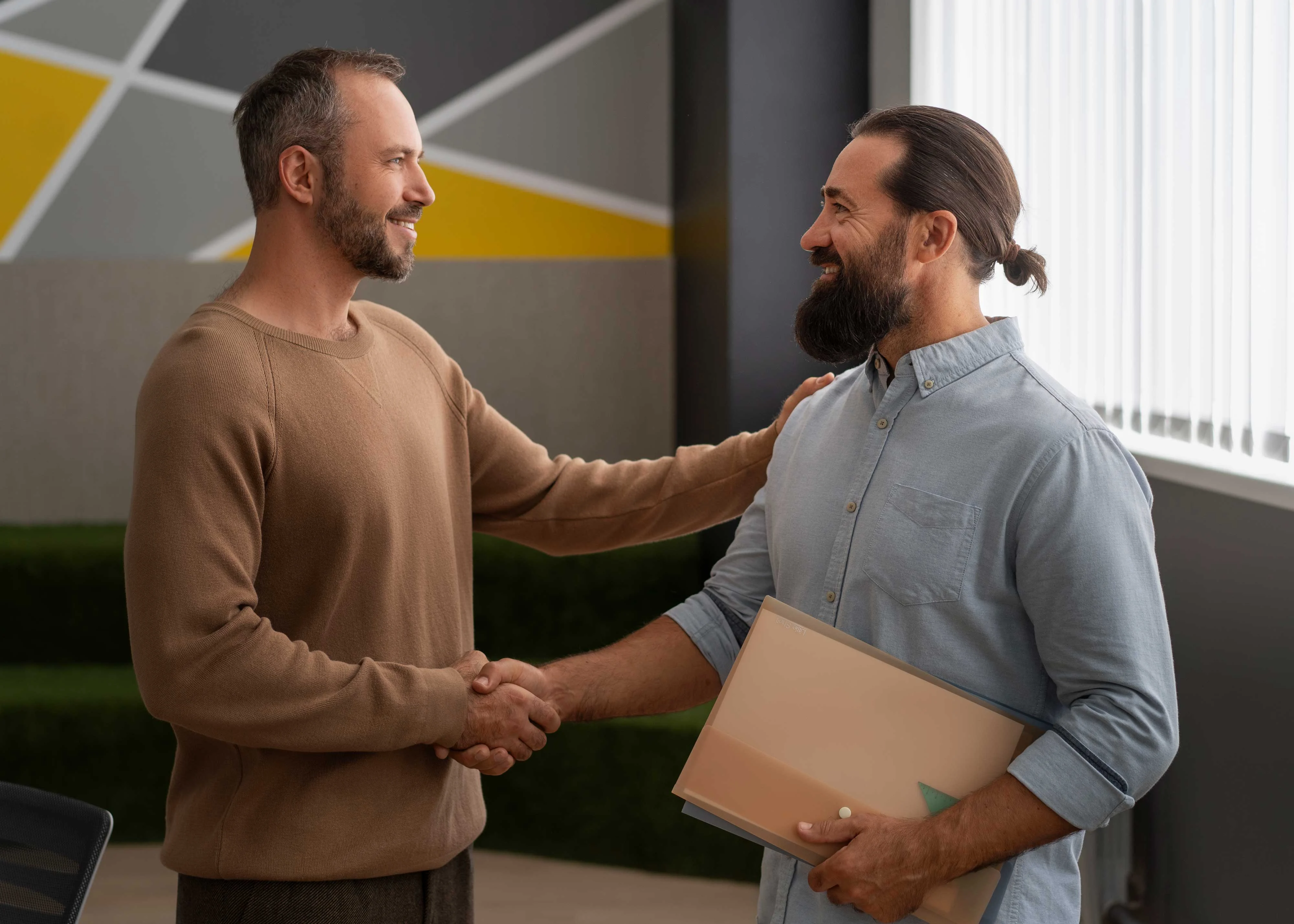 Business Meeting and Collaboration Two men shaking hands in a modern office setting, one is holding a folder.