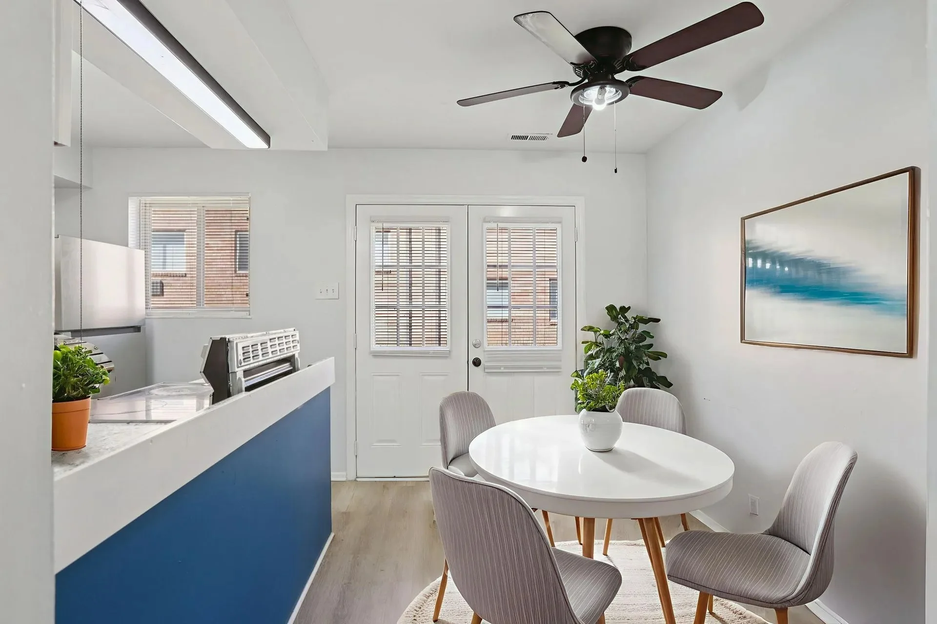 A modern kitchen and dining area featuring a round white table with grey chairs, a potted plant, and a kitchen counter with a blue accent wall.