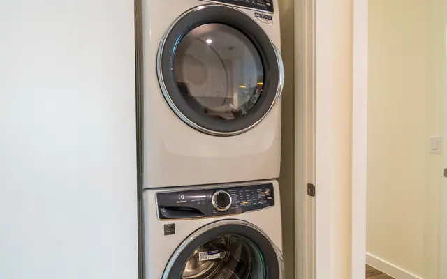 A stacked washer and dryer in a small alcove, with beige walls and wooden flooring.