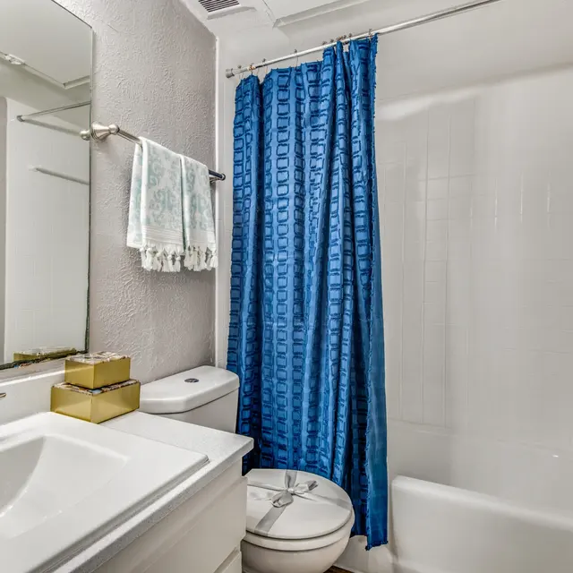 A small bathroom featuring a bathtub with a shower curtain, a toilet, and a sink with a mirror above it. The shower curtain is blue and the walls are light-colored. Shelves with towels and decorative baskets are visible in the background.