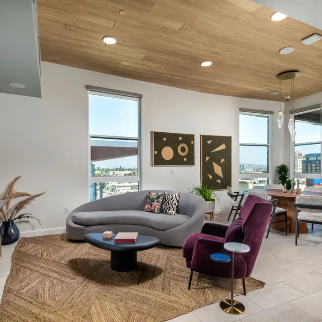 Contemporary Living Room Interior A modern living room featuring a curved gray sofa, a round black coffee table, and a purple armchair. The room has large windows offering a view, abstract artwork on the walls, and a wooden ceiling. There are plants and decorative items in a stylish setting.