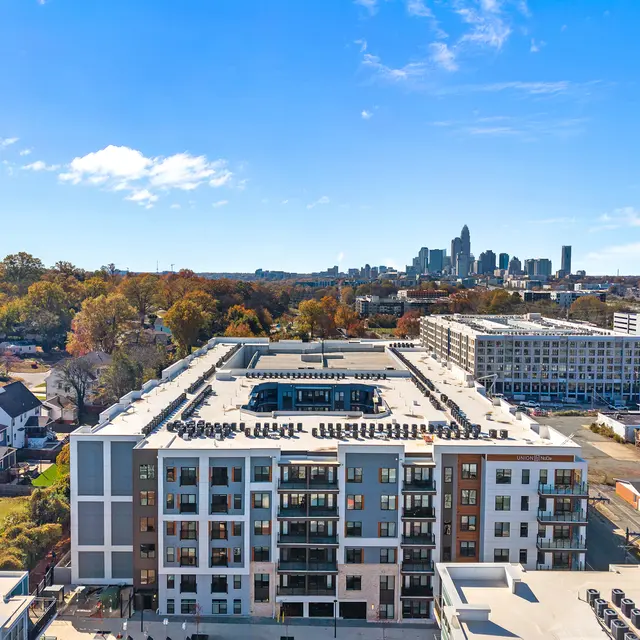 Aerial view of a modern apartment complex with a rooftop garden. In the background, city skyline and autumn trees are visible.