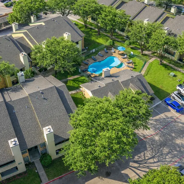 Aerial view of an apartment complex featuring multiple buildings, a swimming pool, and parking spaces surrounded by trees.