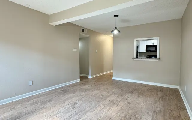 An empty living room featuring light brown wood-style flooring, beige walls, and a kitchen visible through an opening. A pendant light hangs from the ceiling.