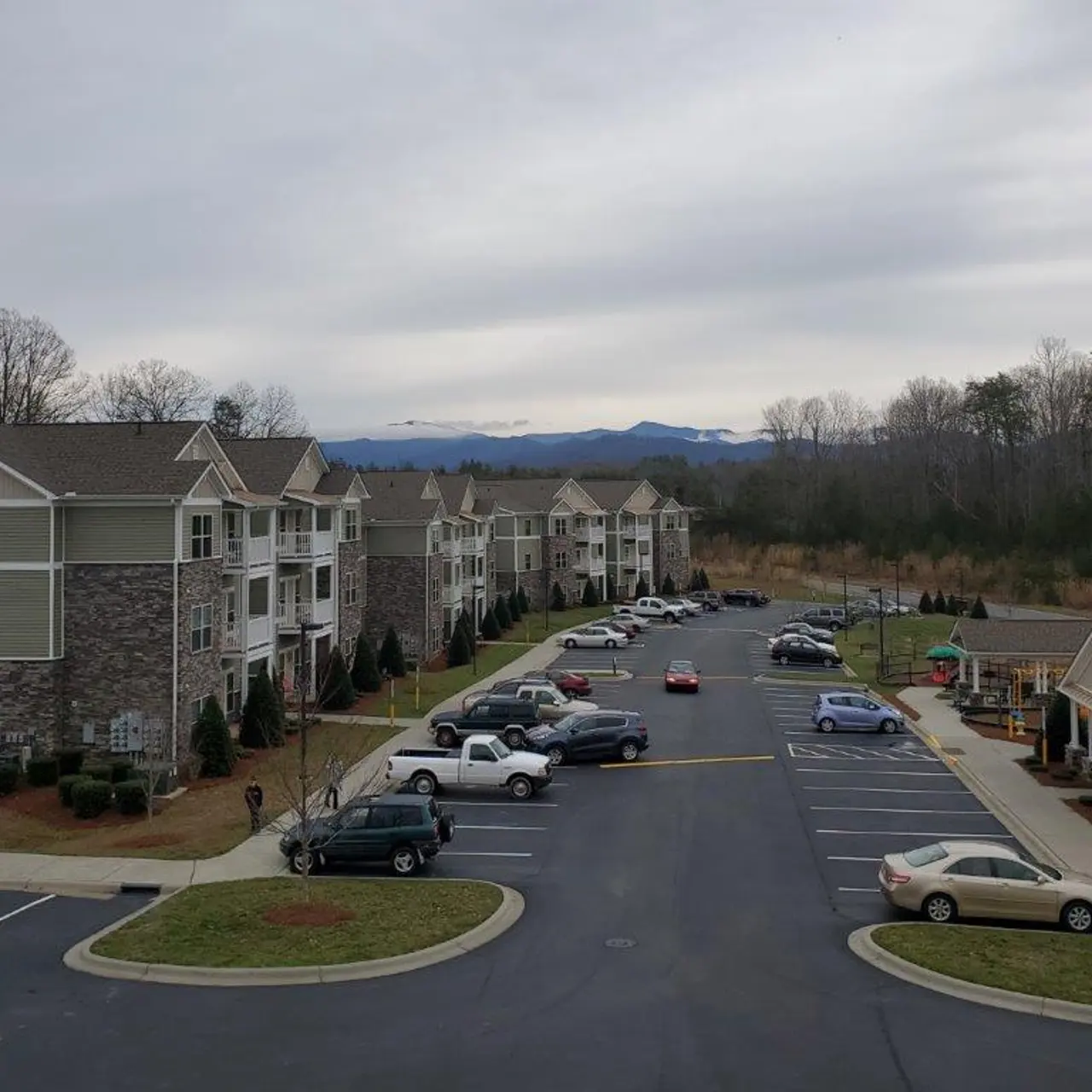 A view of an apartment complex with multiple buildings and a parking lot. There are several cars parked, and in the background, trees and low mountains can be seen under a cloudy sky.