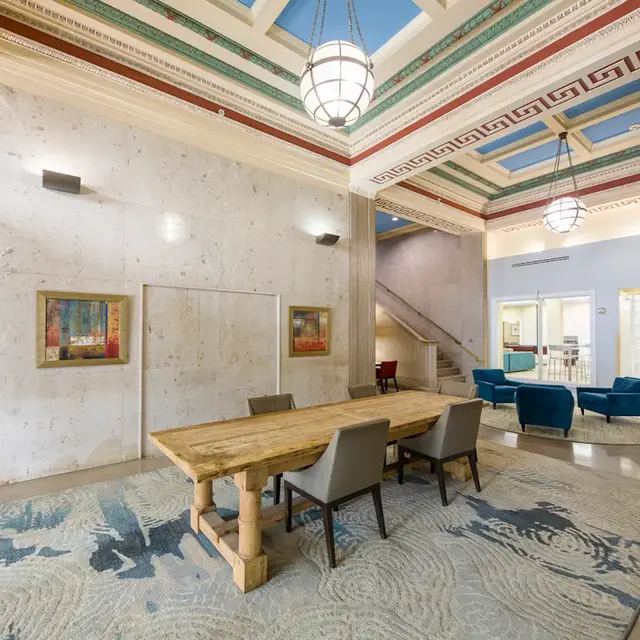 Elegant Communal Lounge Area An elegant communal space featuring a wooden dining table surrounded by chairs, with a decorative rug and artistic wall panels. The ceiling is adorned with intricate designs and light fixtures, while clear glass doors lead to another area.