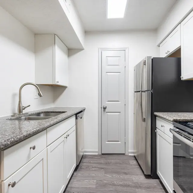 A modern kitchen featuring white cabinetry, granite countertops, stainless steel appliances, and a door leading to another room.
