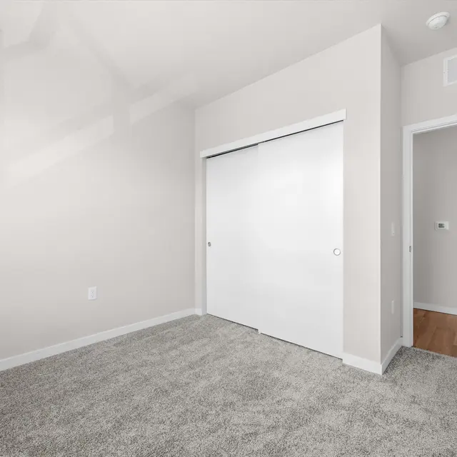 Empty room with light gray carpet and beige walls, featuring sliding doors in the center and a doorway on the right.