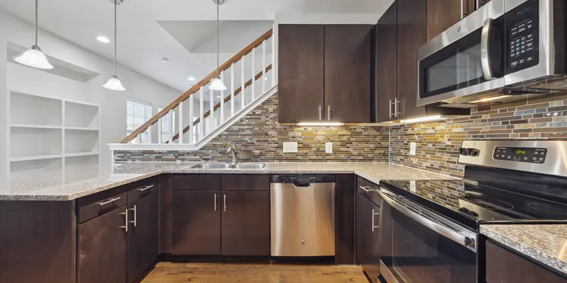 A modern kitchen featuring dark cabinetry, stainless steel appliances, and a tile backsplash with subtle textures. The kitchen island is equipped with a sink, and there are pendant lights above. A staircase is visible in the background, leading to an upper level.