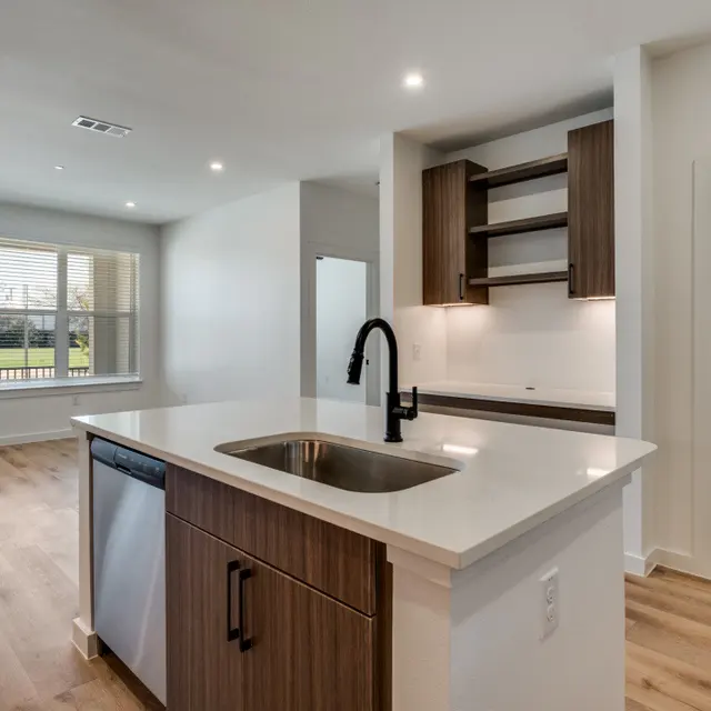 A modern kitchen featuring a white countertop, dark wood cabinetry, and an integrated dishwasher. In the background, there is a laundry area with stacked appliances and a window overlooking a green area.