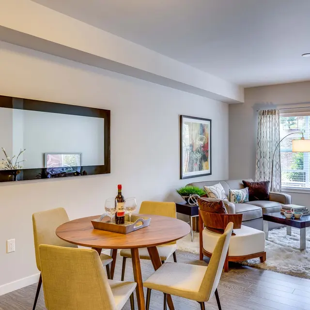 A cozy modern living room with a round dining table and chairs, a comfortable sofa set near a window, and modern decor including a mirror and a piece of wall art.