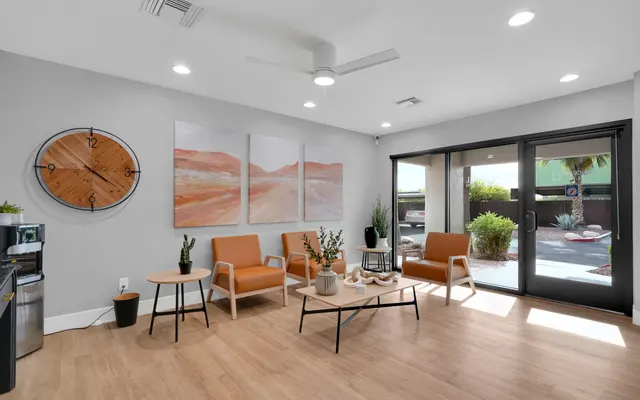 A modern living room featuring two orange chairs, a wooden clock on the wall, and abstract art pieces. Large glass doors lead to an outdoor area with plants.