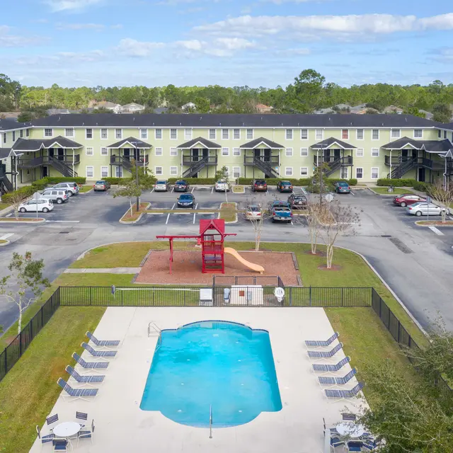 Aerial view of an apartment complex with a swimming pool in the foreground and a playground nearby. The building features multiple floors and balconies.