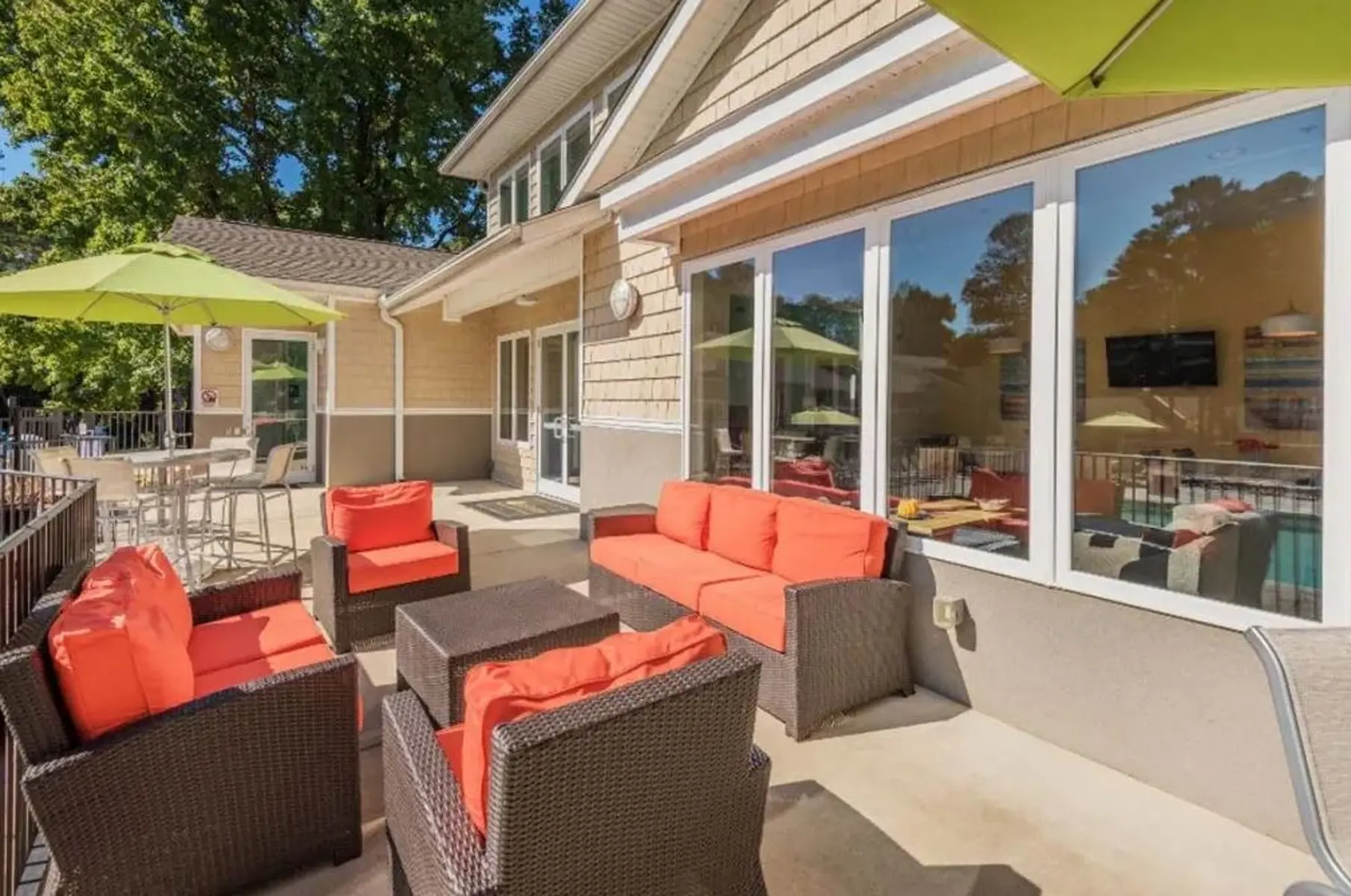 An outdoor lounge area featuring comfortable seating with orange cushions and a dark wicker coffee table. Several green umbrellas provide shade, and there are large windows showcasing a bright interior. The surroundings include trees and a pool area visible through the windows.