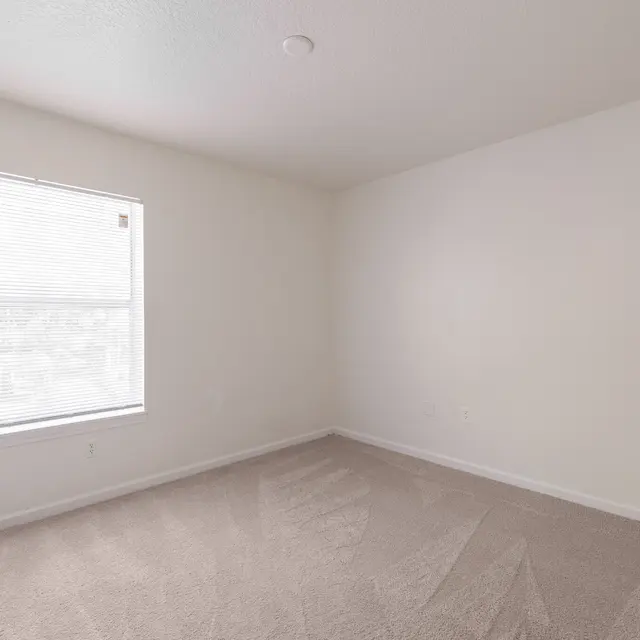 A vacant bedroom featuring light beige walls, a large window with blinds, and plush carpet flooring.