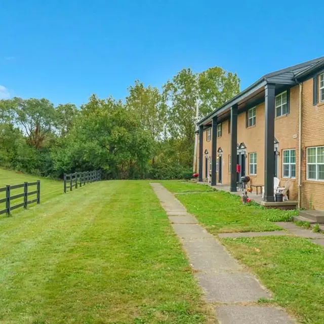 A view of a brick townhouse complex with a walkway and grassy area. There are trees in the background and a fence along the side.