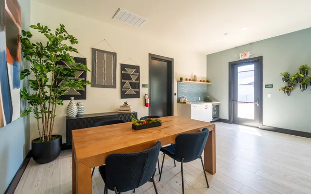 A modern dining area featuring a wooden table with black chairs, plants in pots, and wall decorations. The room has a light color scheme with natural light coming through a door.