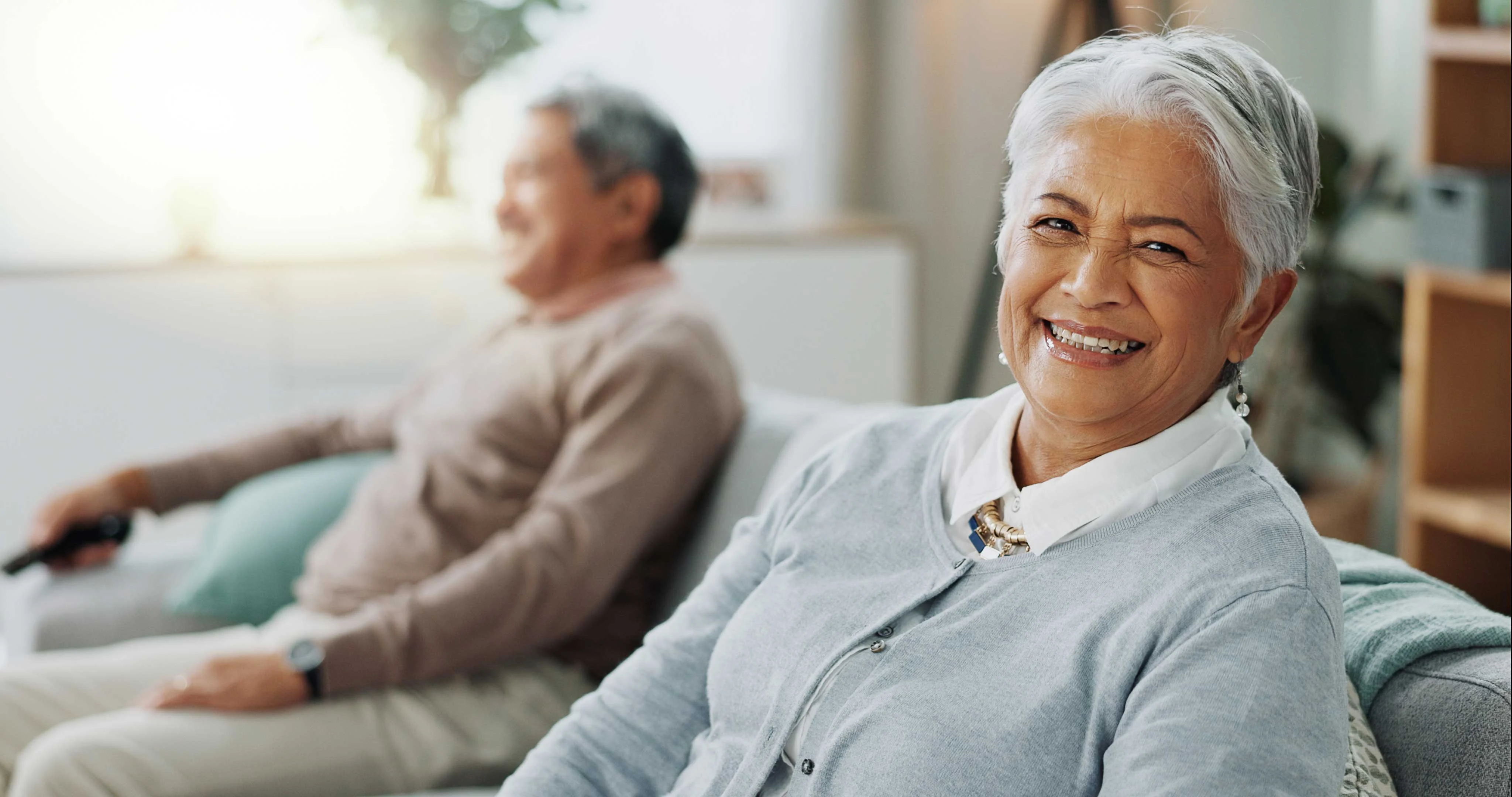 A senior woman smiling at the camera while sitting in a living room. Behind her, a senior man is seated on a couch, looking relaxed. The room has natural light and a cozy atmosphere.