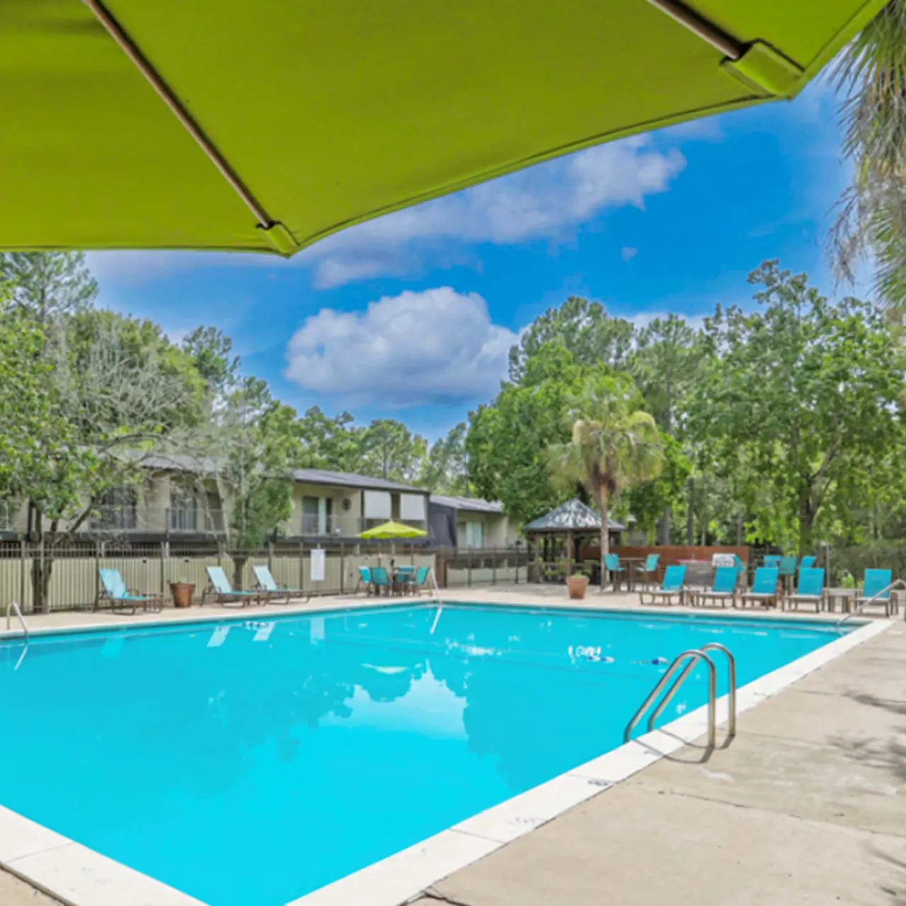 A spacious outdoor swimming pool surrounded by lounge chairs and palm trees, with an umbrella providing shade.