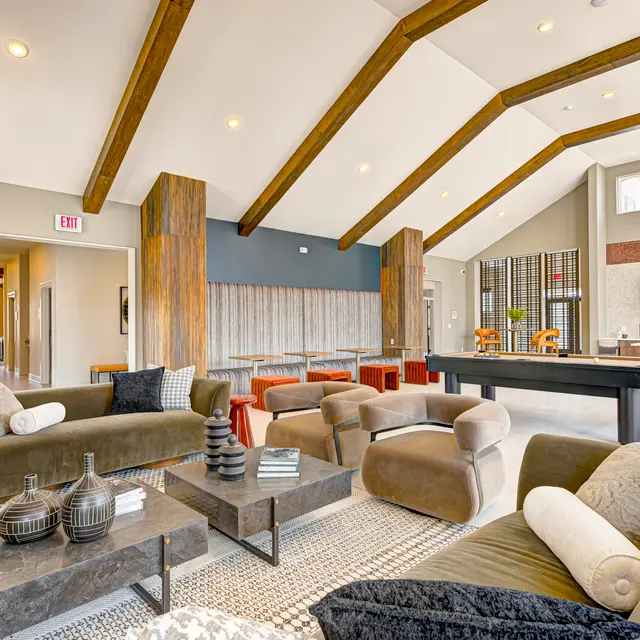 A spacious and stylish living room featuring high ceilings with wooden beams, modern furniture including sofas and coffee tables, and a dining area in the background. Large windows allow natural light to fill the space.