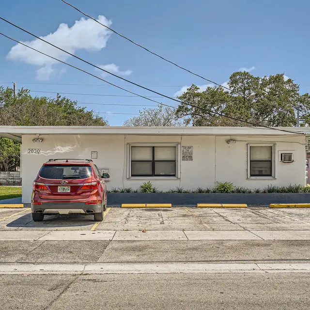 A low-profile building featuring a flat roof, with a red SUV parked in front. The facade has several windows and is surrounded by green grass and trees. The scene is under a clear blue sky.