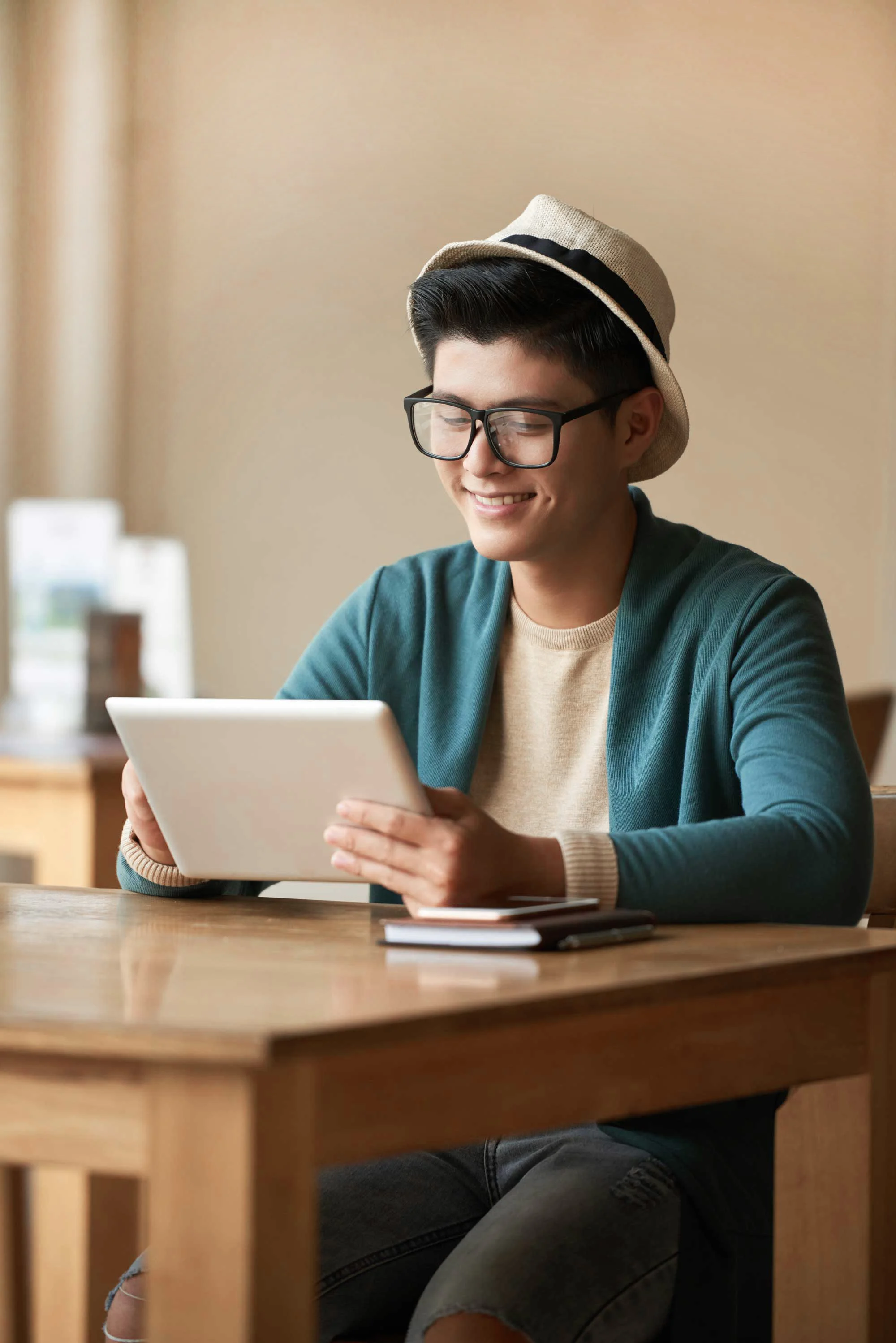 A young man sitting at a wooden table in a café, smiling while looking at a tablet. He is wearing a light-colored hat, glasses, and a blue cardigan, with a notepad and a smartphone on the table beside him. The background is softly blurred, giving a warm and inviting atmosphere.