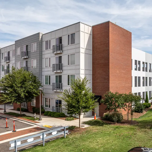 A modern multi-story apartment building featuring a mix of brick and sleek metal siding. Surrounding lawns and trees provide greenery, while construction equipment is visible nearby.
