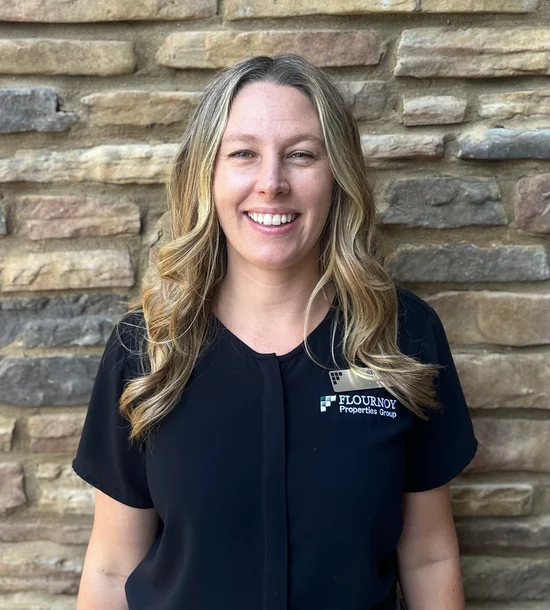 A smiling woman with long, wavy hair, wearing a black shirt with the logo of a real estate company, standing in front of a stone wall.