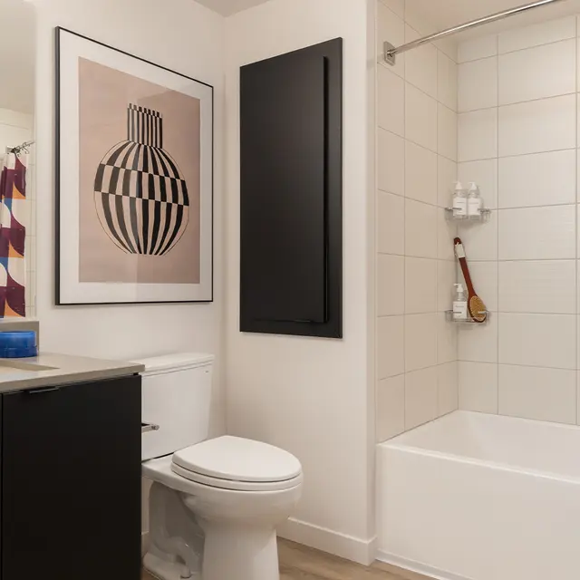 Interior view of a modern bathroom featuring a bathtub, toilet, and sink. The walls are tiled with a large abstract art piece on display, complemented by colorful shower curtain patterns.